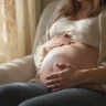 Femme enceinte en fin de grossesse assise confortablement, main posée sur le ventre, atmosphère rassurante